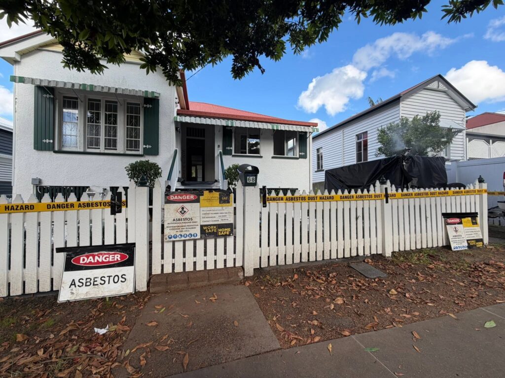 White picket fence around a house with yellow 'DANGER ASBESTOS' tape and warning signs; construction hazard area.