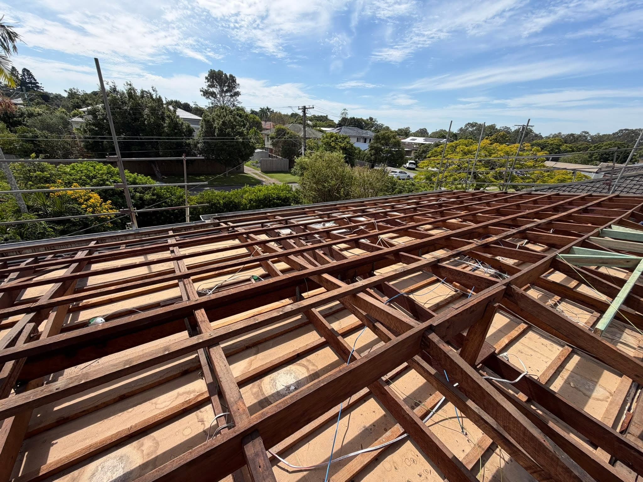 Exposed wooden roof framing on a construction site with a suburban street and trees in the background.