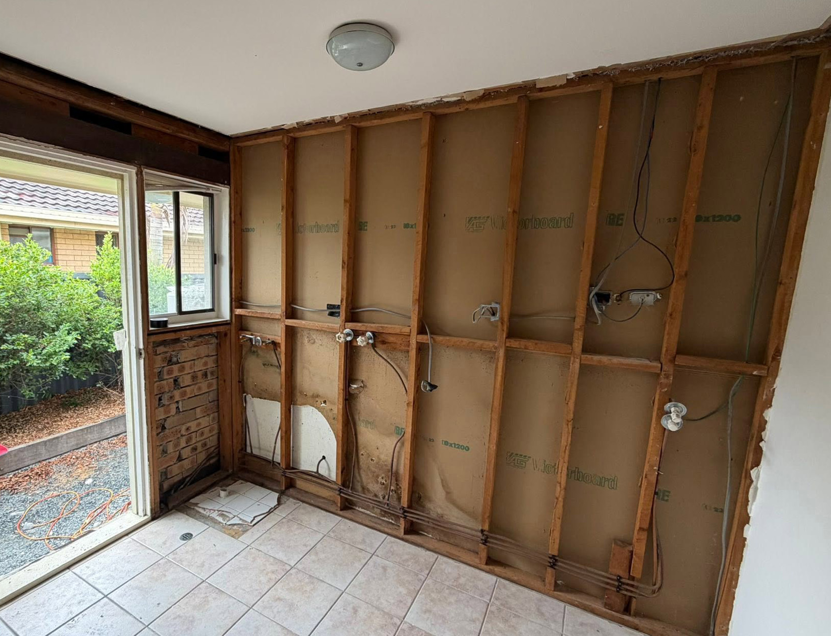 Partially demolished kitchen with exposed studs, plumbing and wiring along the wall; door to outside and a window visible.