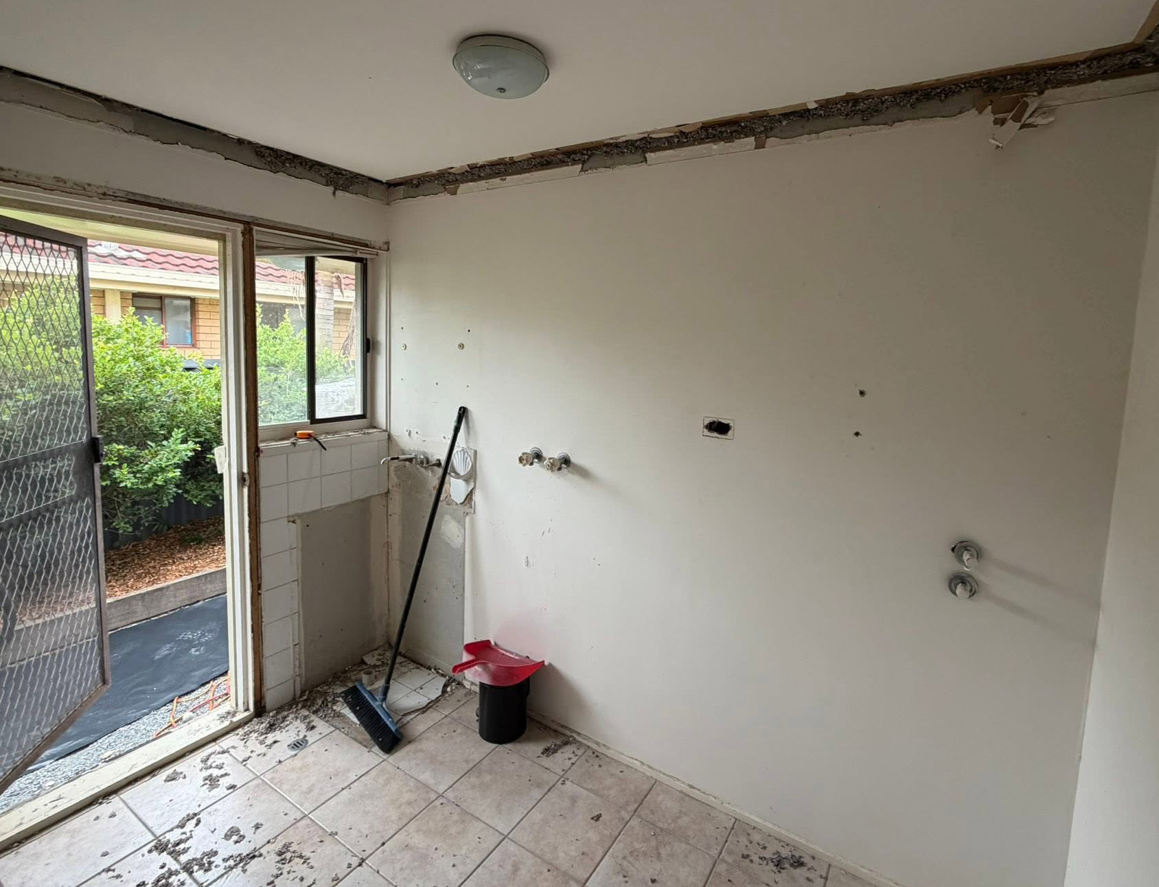Unfinished utility/kitchen area with peeling ceiling, exposed pipes, and wall holes; cleaning tools and a red dustpan on the floor near a sliding door to a garden courtyard