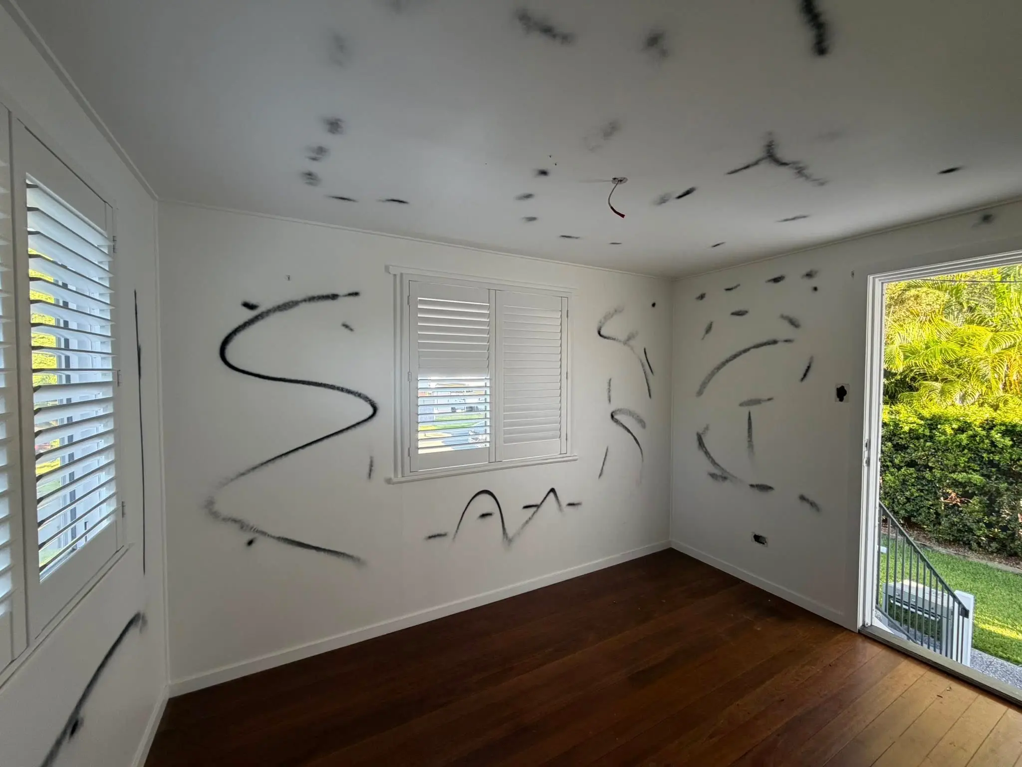 White room with random black graffiti scribbles on walls and ceiling; window blinds on the left and a large door with greenery outside.