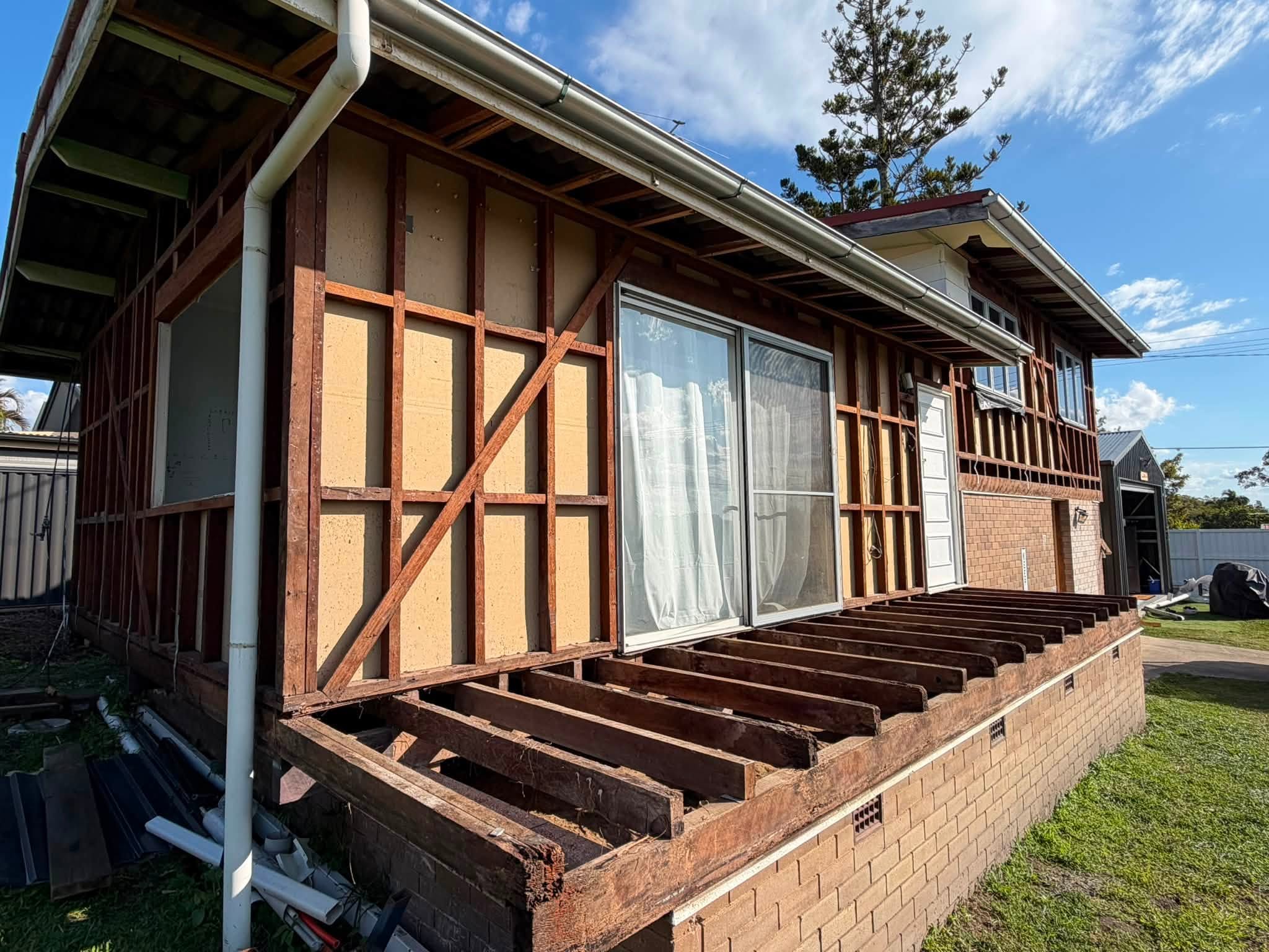Side view of a house under renovation with exposed wooden wall framing and a partially built deck frame, white gutters along the roof, and large sliding glass doors under a blue sky