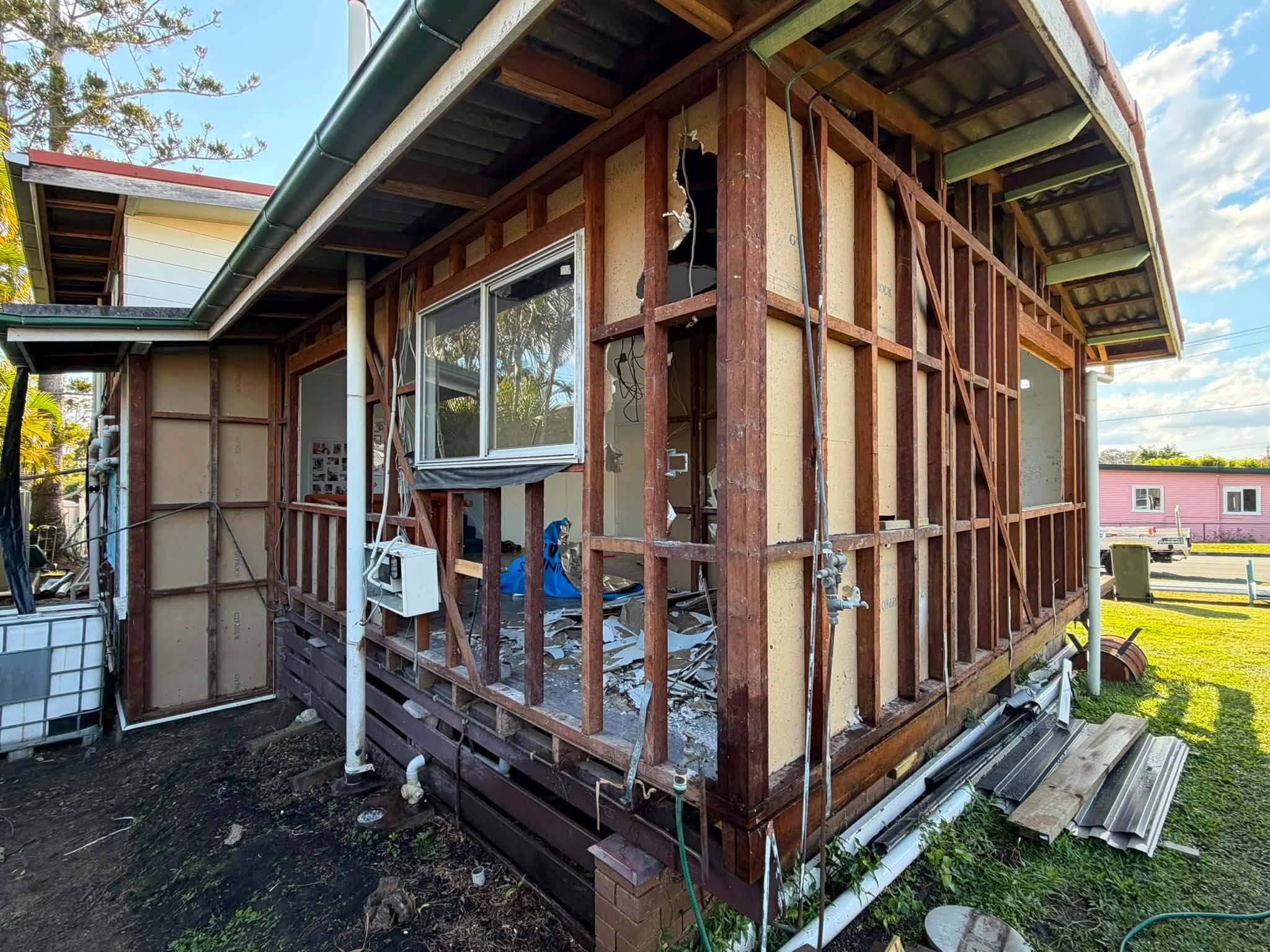 Exterior of a house under renovation with exposed wooden studs, missing siding, and scattered debris outside the unfinished wall.