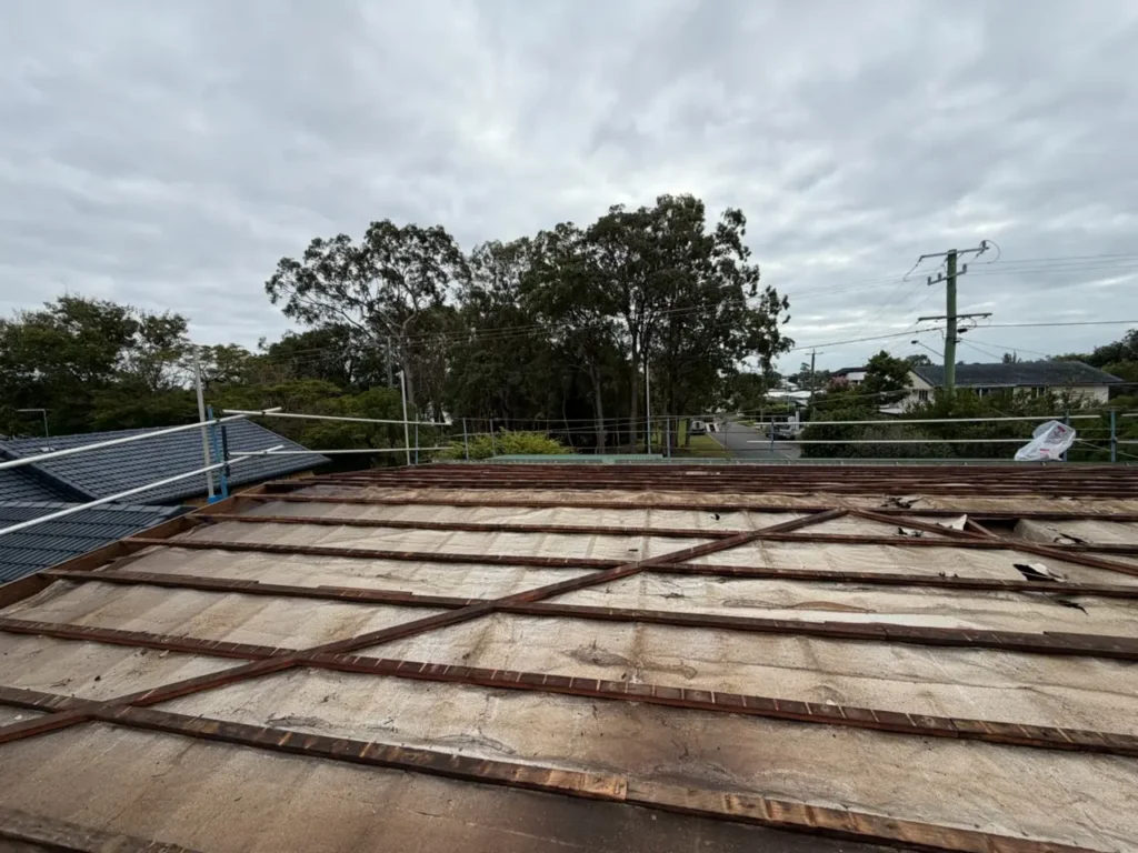 Rooftop under construction with metal battens and safety railing, overlooking a suburban street and trees under a cloudy sky.