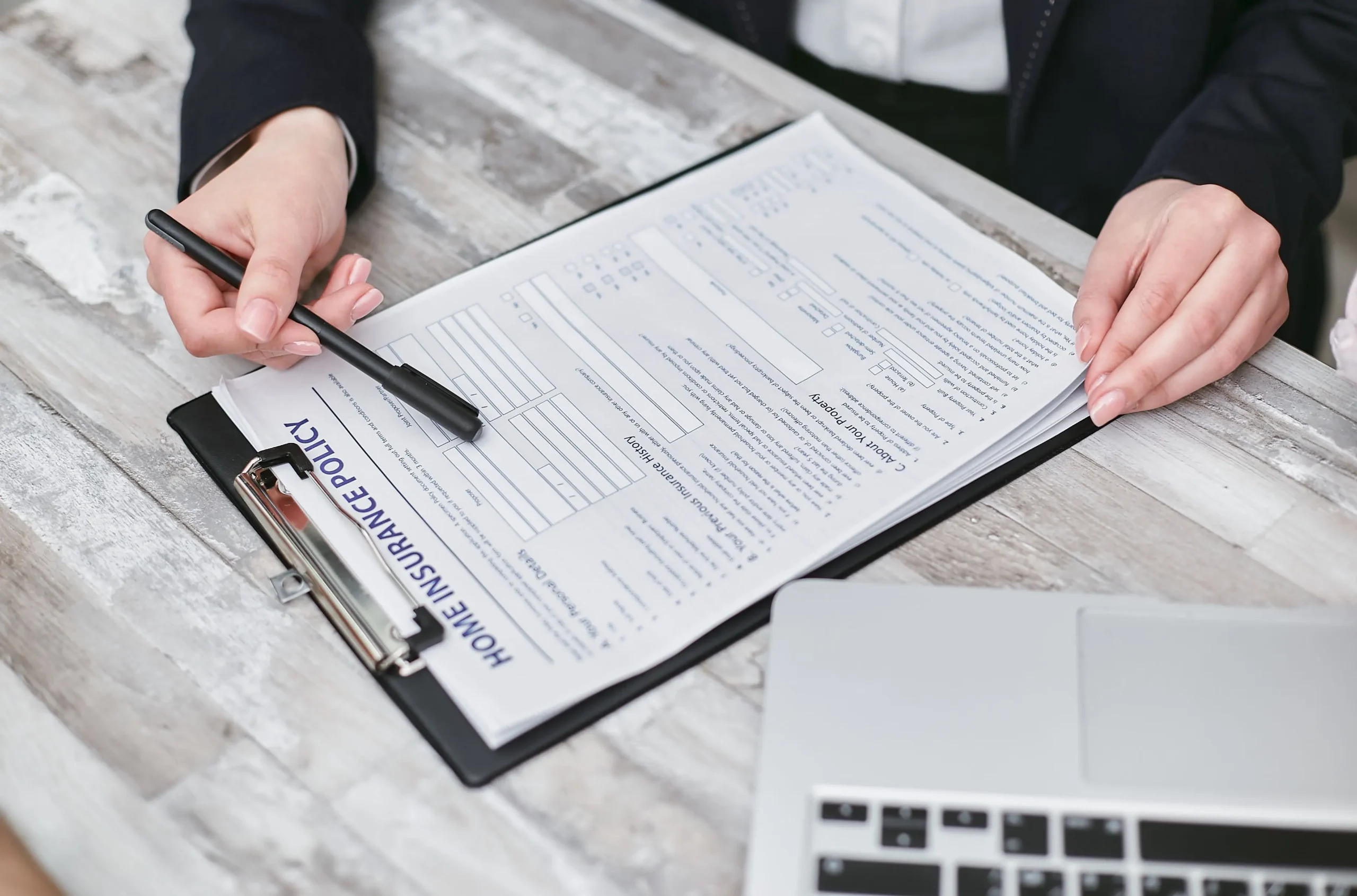Person’s hands fill out a form on a clipboard with a black pen nearby a laptop on a wooden desk